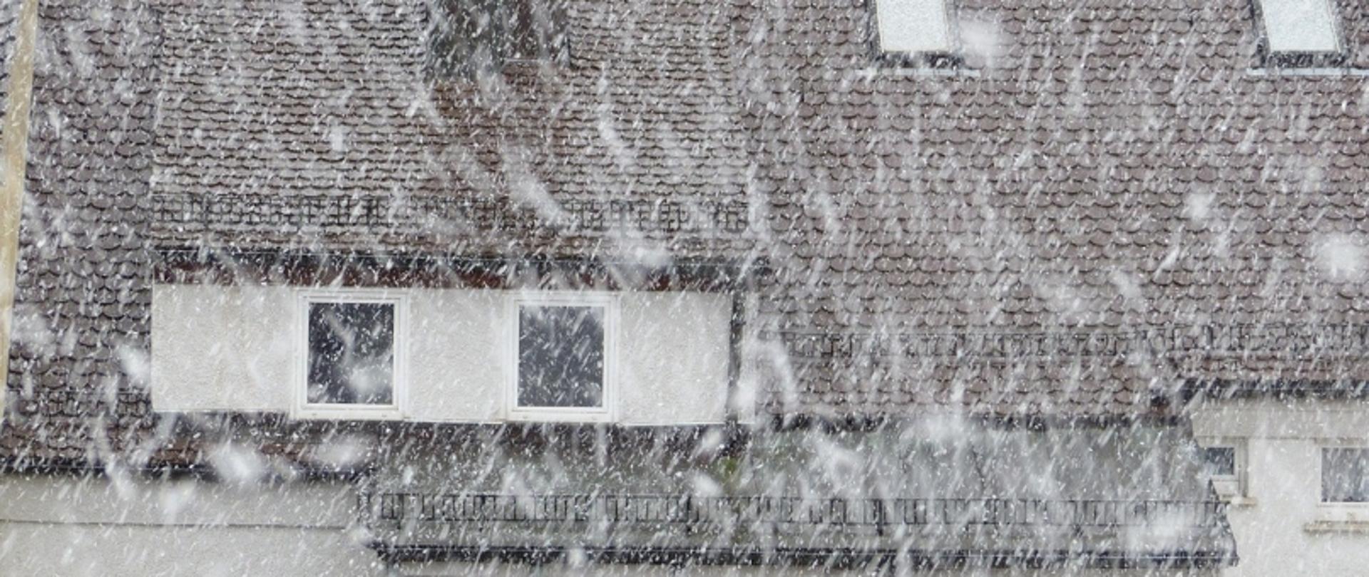 Budynek mieszkalny podczas intensywnych opadów śniegu, z zaśnieżonym dachem i balkonem – ilustracja ostrzeżenia pogodowego.