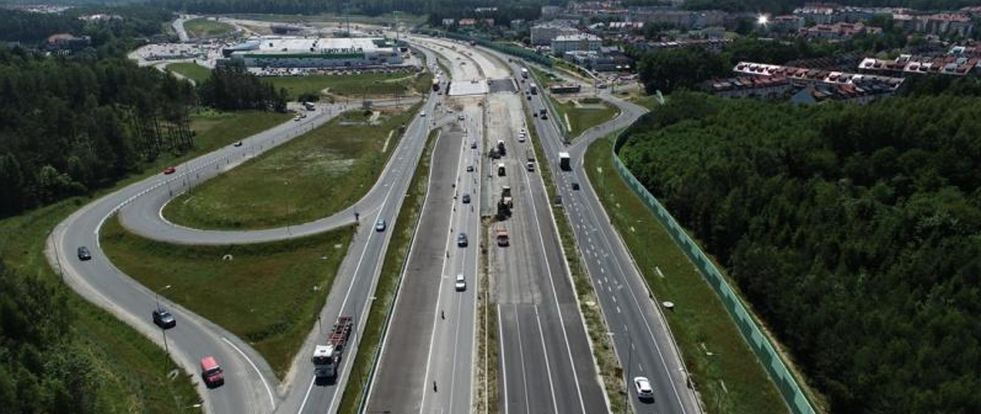 Zdjęcie przedstawia budowę węzła Gdynia Wielki Kack na Obwodnicy Trójmiasta. W centrum zdjęcia widać trwające prace przy wykonaniu nawierzchni bitumicznej, po bokach odbywa się ruch samochodów.