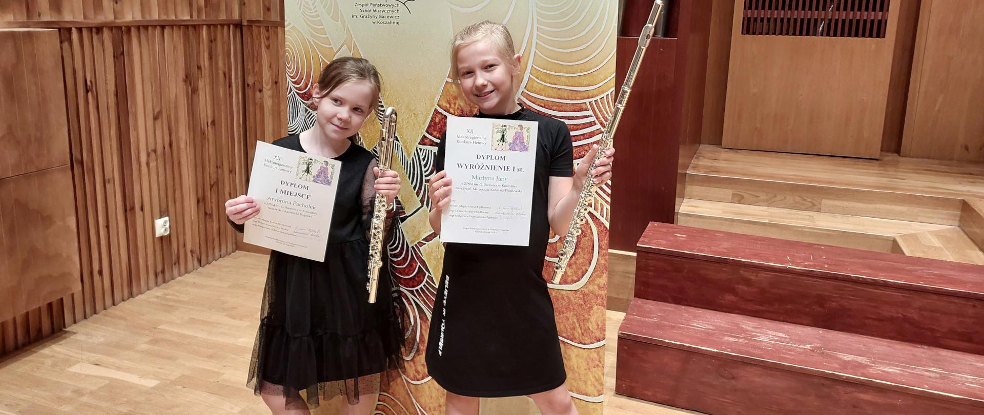 Uczennice ZPSM w ręku trzymają flet poprzeczny i dyplom, na tle schodów drewnianych i banera