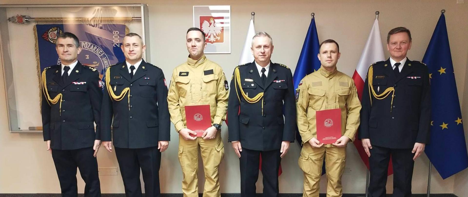 Zdjęcie przedstawia sześciu mężczyzn w mundurach strażackich, stojących obok siebie w pomieszczeniu biurowym podczas uroczystości.
W tle, za mężczyznami, widać białą ścianę, na której wiszą cztery flagi. Są to prawdopodobnie flagi Polski oraz Unii Europejskiej, ustawione naprzemiennie. Po lewej stronie zdjęcia, na ścianie, znajduje się także godło lub tablica z napisem "Państwowa Straż Pożarna". Podłoga jest wyłożona szarym dywanem. Oświetlenie jest jasne i równomierne.