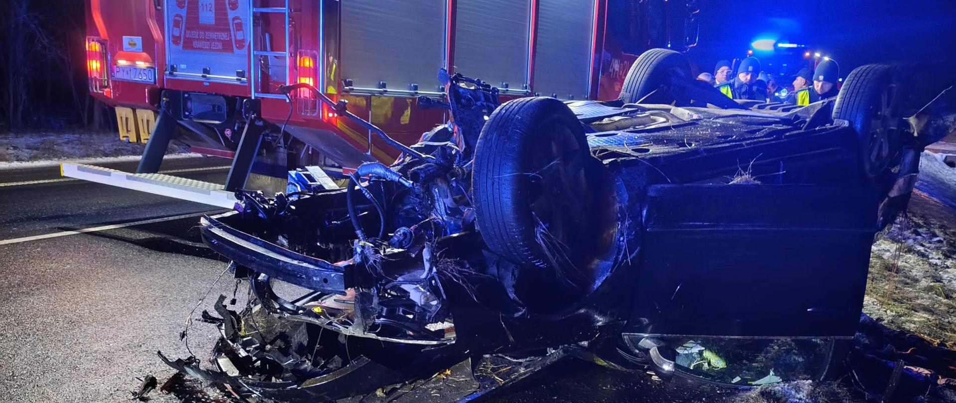 Na zdjęciu widoczny jest wypadek drogowy – samochód osobowy leży na dachu na jezdni, z poważnie uszkodzonym przodem i rozrzuconymi elementami karoserii. Obok znajduje się samochód straży pożarnej z włączonym oświetleniem ostrzegawczym, a w tle widać strażaków oraz pojazdy służb ratunkowych zabezpieczające miejsce zdarzenia.