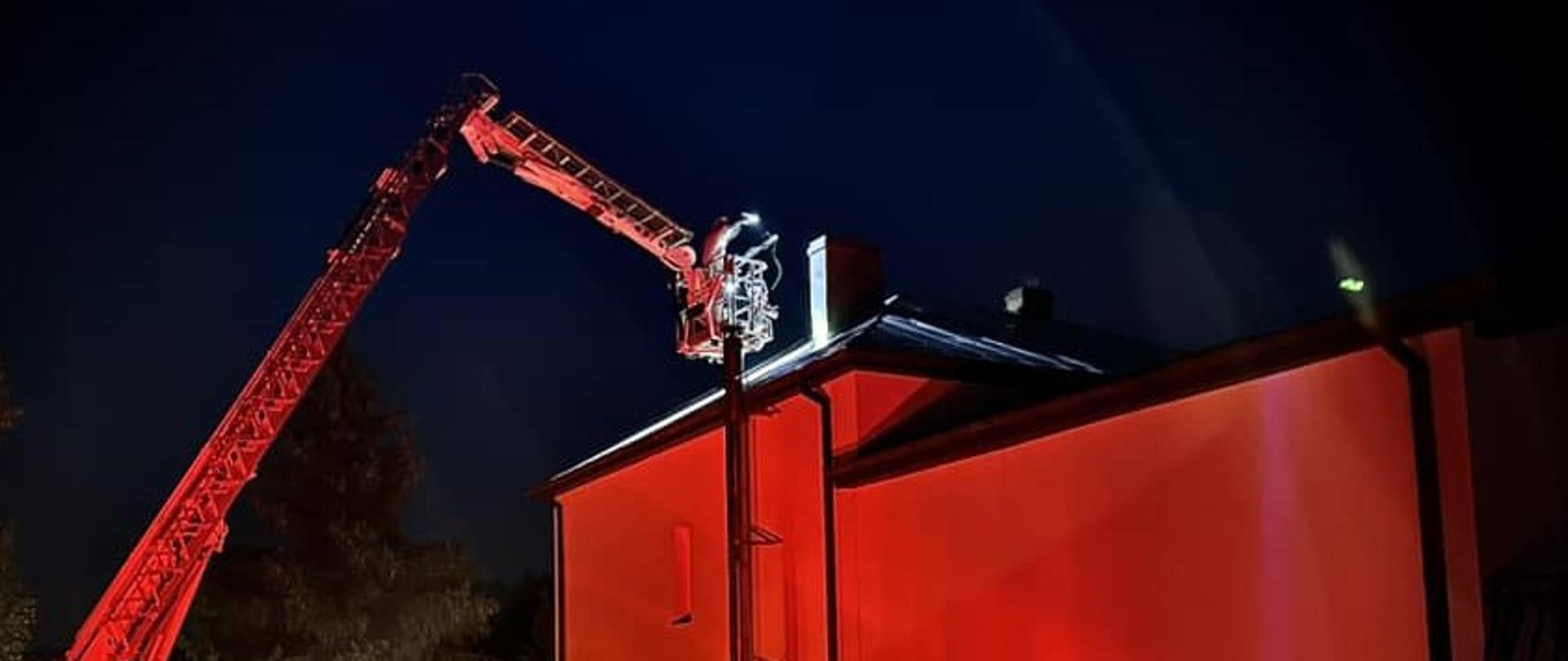 Zdjęcie przedstawia scenę nocnej akcji ratowniczej, gdzie wóz strażacki z wysuniętą drabiną z koszem jest używany do działań przy budynku. Drabina jest uniesiona i skierowana w stronę dachu budynku, gdzie znajduje się strażak w koszu. Budynek, o jasnych ścianach, jest silnie oświetlony czerwonym światłem, co może sugerować syreny pojazdu strażackiego lub specjalne oświetlenie używane podczas akcji. W tle widać ciemne drzewa i nocne niebo. Na tyle wozu strażackiego widoczny jest numer "601-53".