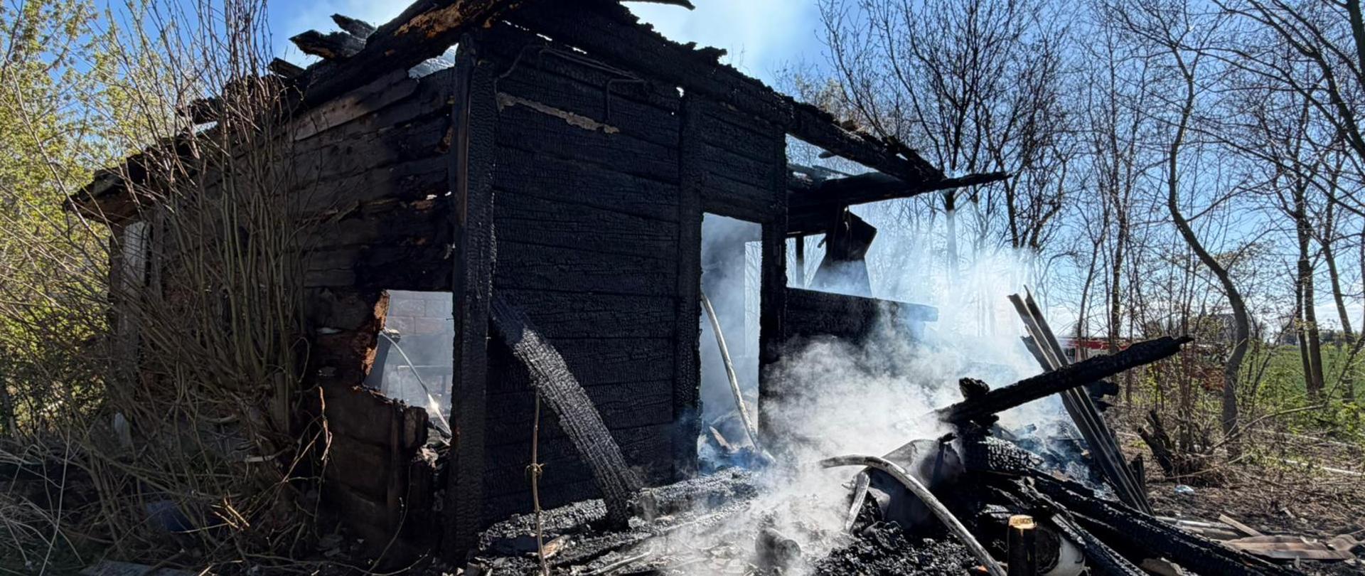 Zgliszcza niewielkiego drewnianego budynku po pożarze. Ściany i dach są poważnie zniszczone i zwęglone, część konstrukcji zawaliła się. Na pierwszym planie widać stertę spalonych desek i metalowych elementów, z których unosi się dym. W tle stoją nagie drzewa, a nad ruinami widoczne jest jasne niebo.