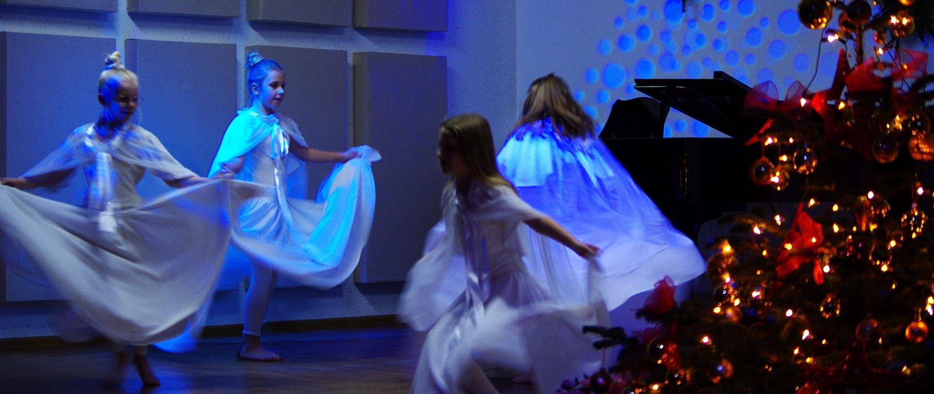 Zdjęcie przedstawia czwrókę dziewczynek w białych sukniach tańczących na scenie szkoły. Scenę oświetla niebieskie, zimne swiatło, z prawej strony stoi ubrana choinka, a w tyle fortepian. Na tylnej ścianie sceny wyświetlają się niebieskie kółka.