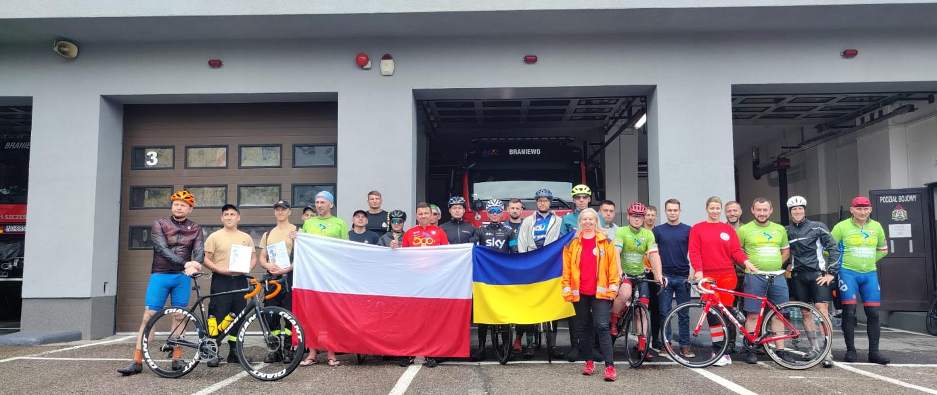 Na zdjęciu na tle garaży bojowych straży pożarnej, uczestnicy rajdu rowerowego. Na wprost stoi kilkudziesięciu uczestników rajdu, kolorowo ubrani, widać rowery w rekach trzymają flagę Polski i Ukrainy.