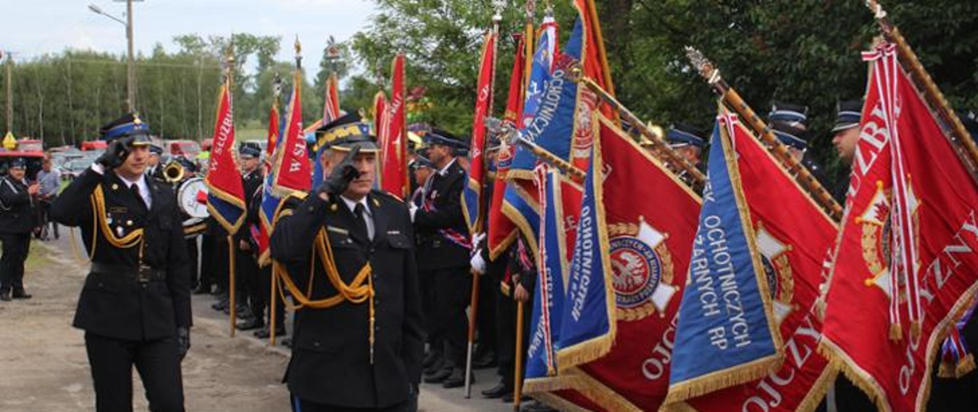 wzdłuż pocztów sztandarowych idzie dwóch salutujących strażaków . strazacy w ciemnych mundurach ze sznurem i w rogatywkach