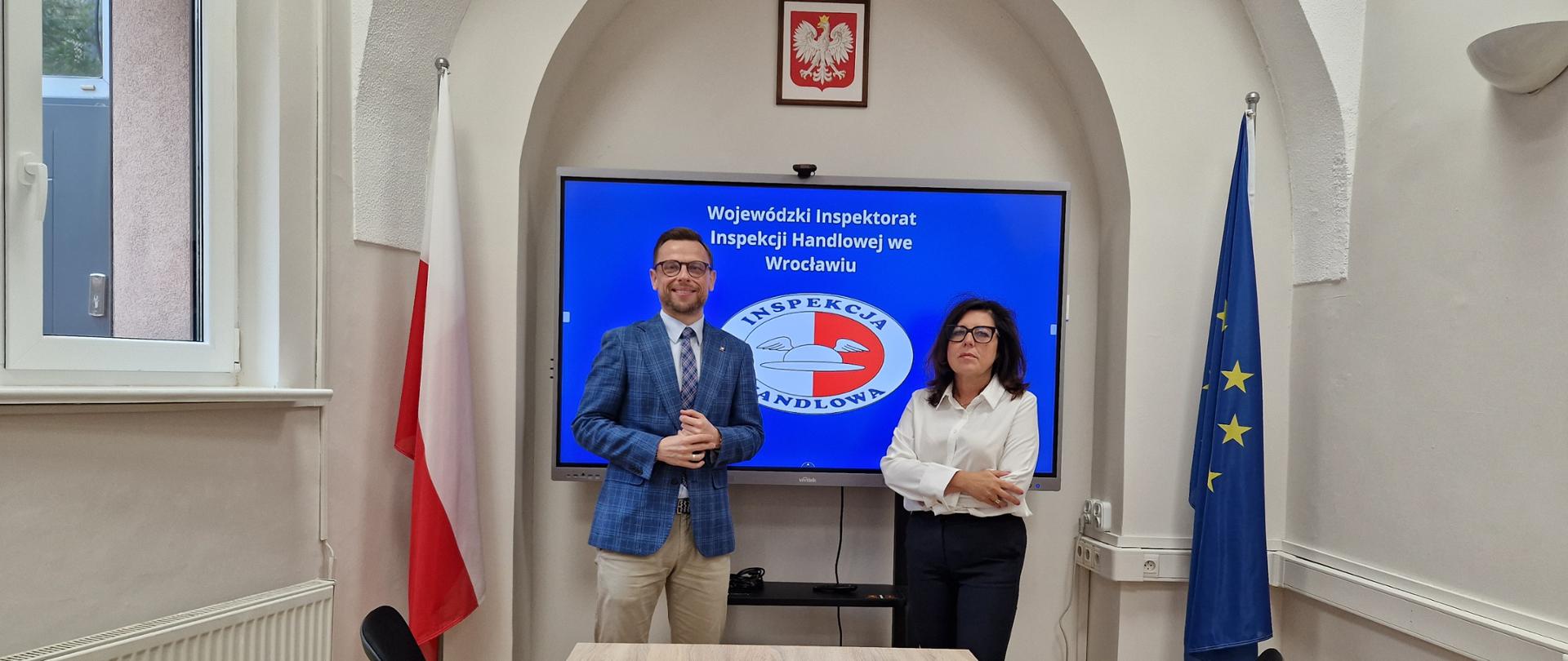Dolnośląski Wojewódzki Inspektor Inspekcji Handlowej i Łódzki Wojewódzki Inspektor Inspekcji Handlowej podczas spotkania we Wrocławiu. W tle tablica multimedialna z logo Inspekcji Handlowej po lewej stronie flaga Polski po prawej stronie flaga UE.
