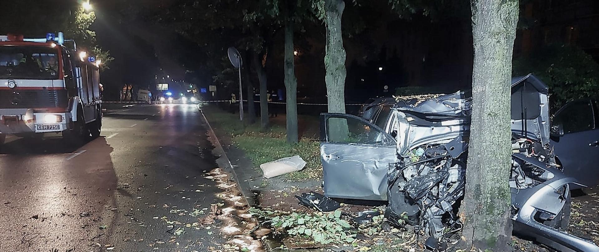 Zdjęcie przedstawia perspektywę ulicy 1-go Maja gdzie miało miejsce zdarzenie. Po prawej stroni e, między drzewami widać zniszczony samochód osobowy, po lewej, na drodze stoi samochód pożarniczy.