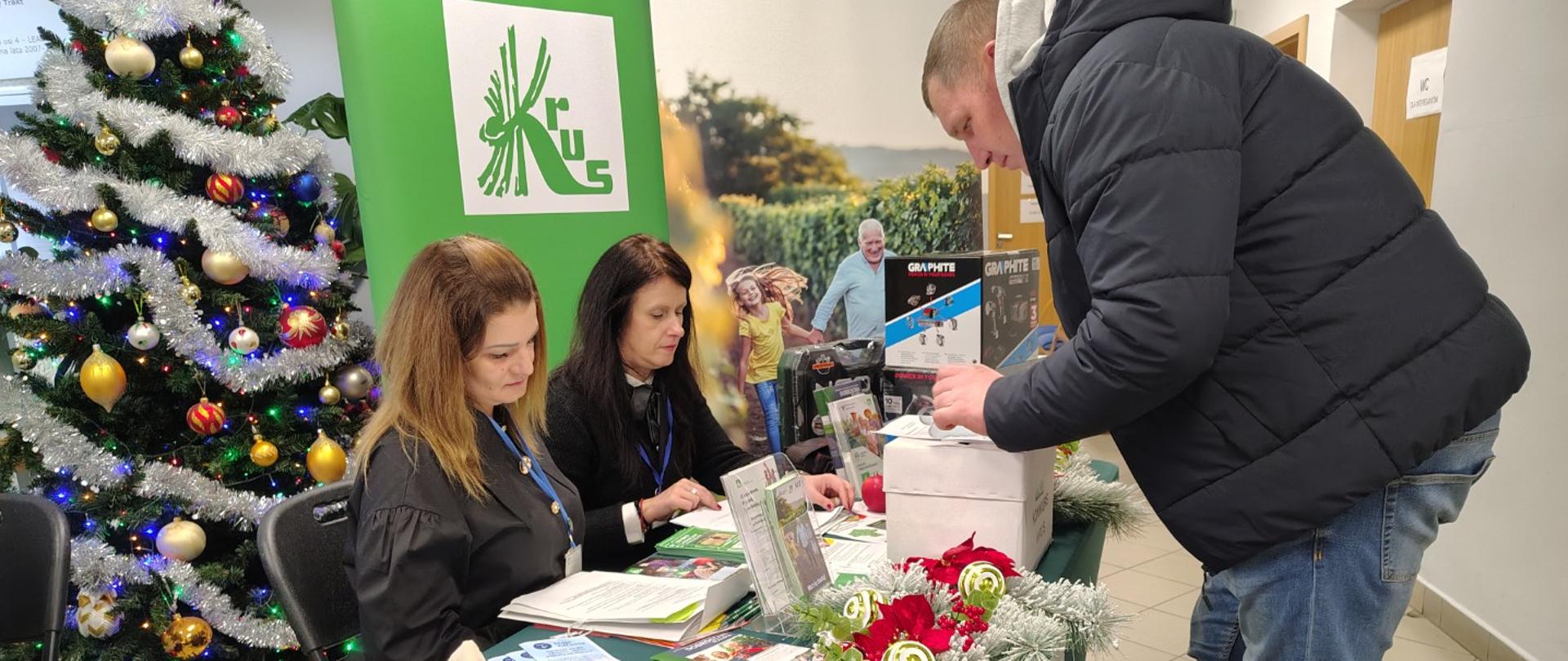 Przedstawicielki KRUS siedzą przy stole, który jest nakryty zielonym obrusem z logo KRUS i sprawdzają testy konkursowe. Przed nimi stoi mężczyzna, który rozwiązuje test wiedzy. Po lewej stronie znajduje się piękna zielona choinka z kolorowymi bombkami. Po jej prawej stronie stoją dwa banery: jeden z logo KRUS, drugi dotyczący profilaktyki badań NFZ.