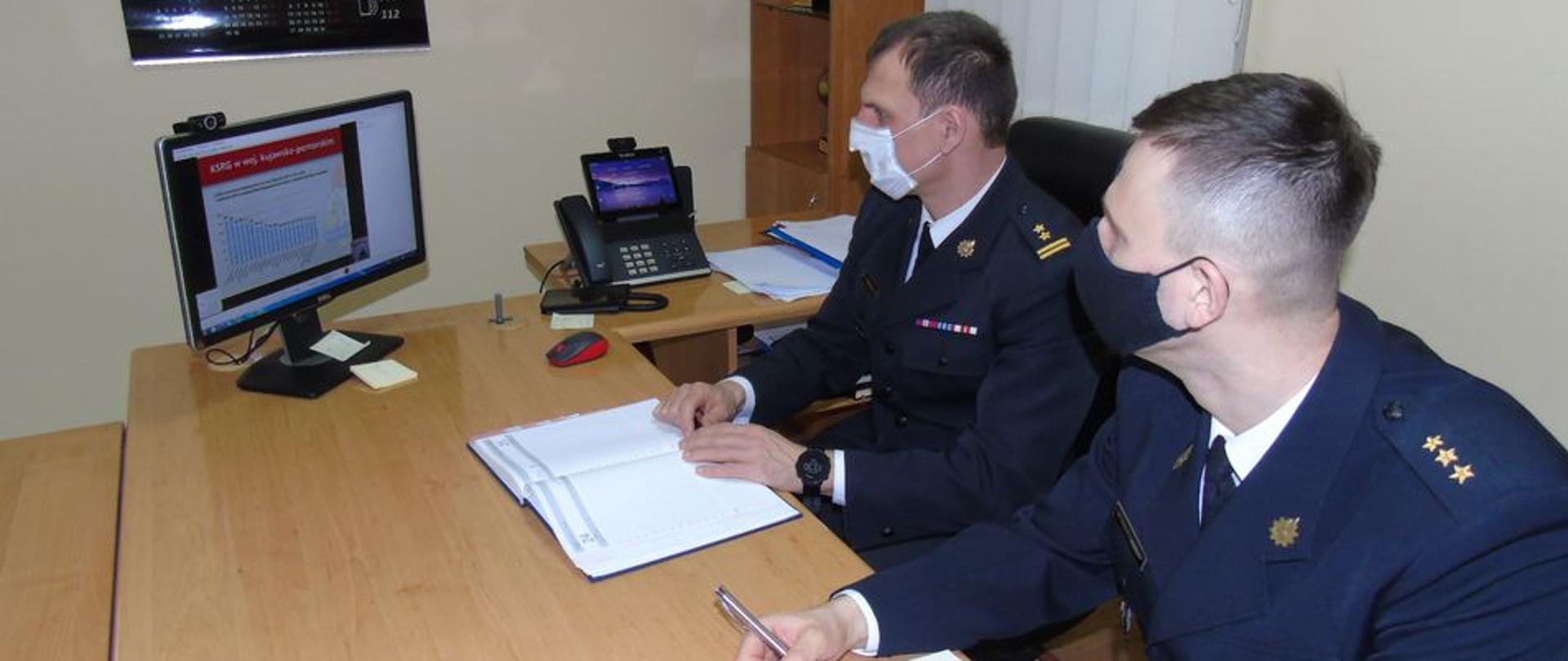 W pomieszczeniu biurowym przy jasnym stole siedzą dwaj strażacy ubrani w czarne mundury wyjściowe ( gabardyna). Na twarzy maja założone maseczki, na stole stoi monitor komputerowy, ich wzrok skierowany jest na monitor, przysłuchują się konferencji.