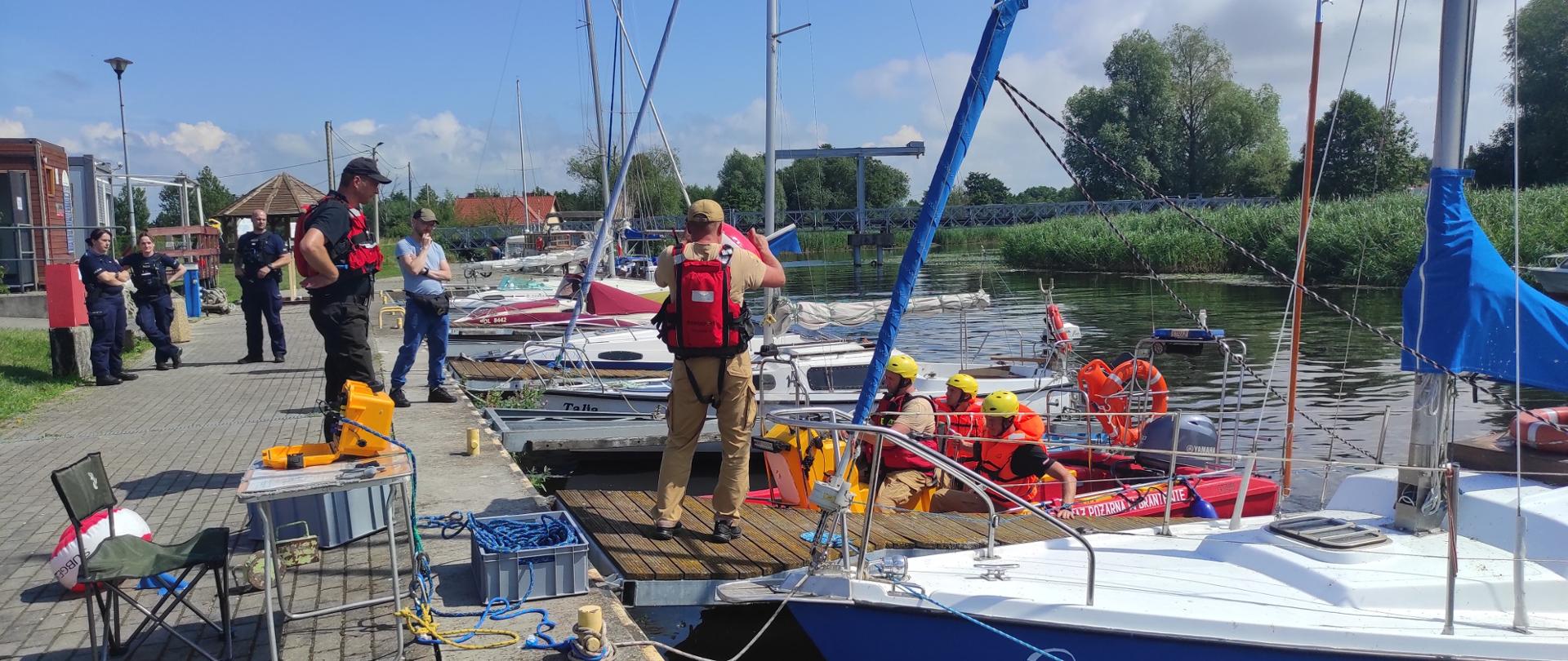 Przystań żeglarska. Na przystania zacumowane jachty. Z prawej strony widać czerwona łódź motorową ze strażakami w żółtych hełmach. Z lewej strony stoi czterech Policjantów. Słonecznie.