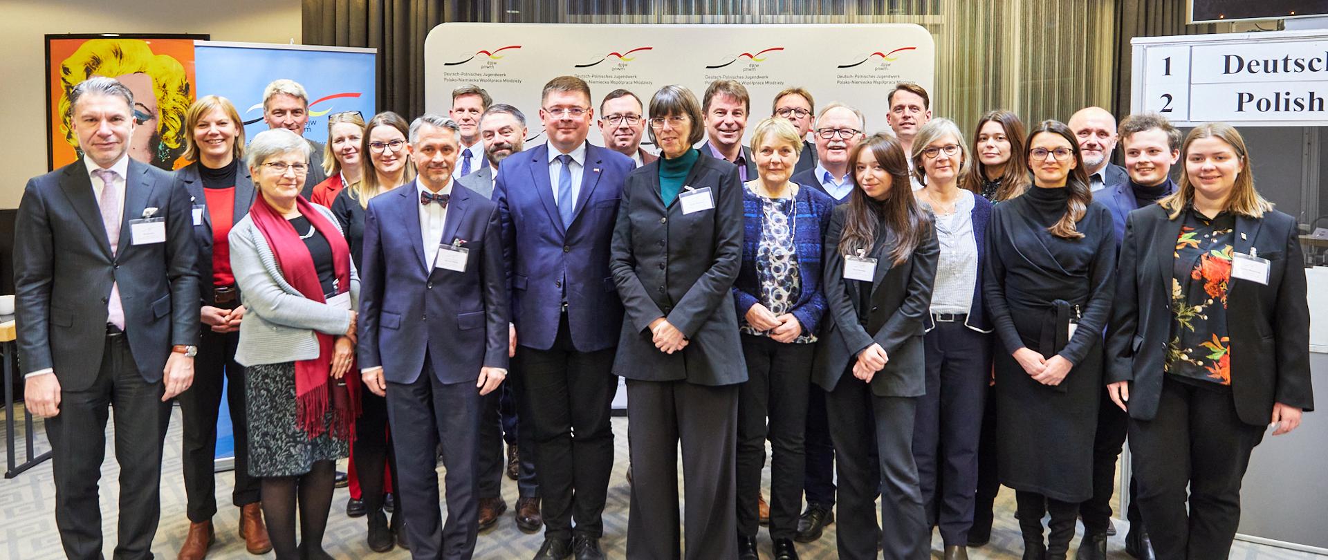 Zdjęcie zbiorowe, uśmiechnięci uczestnicy spotkania stoją w rzędzie pod zasłoniętą zasłonami ścianą.