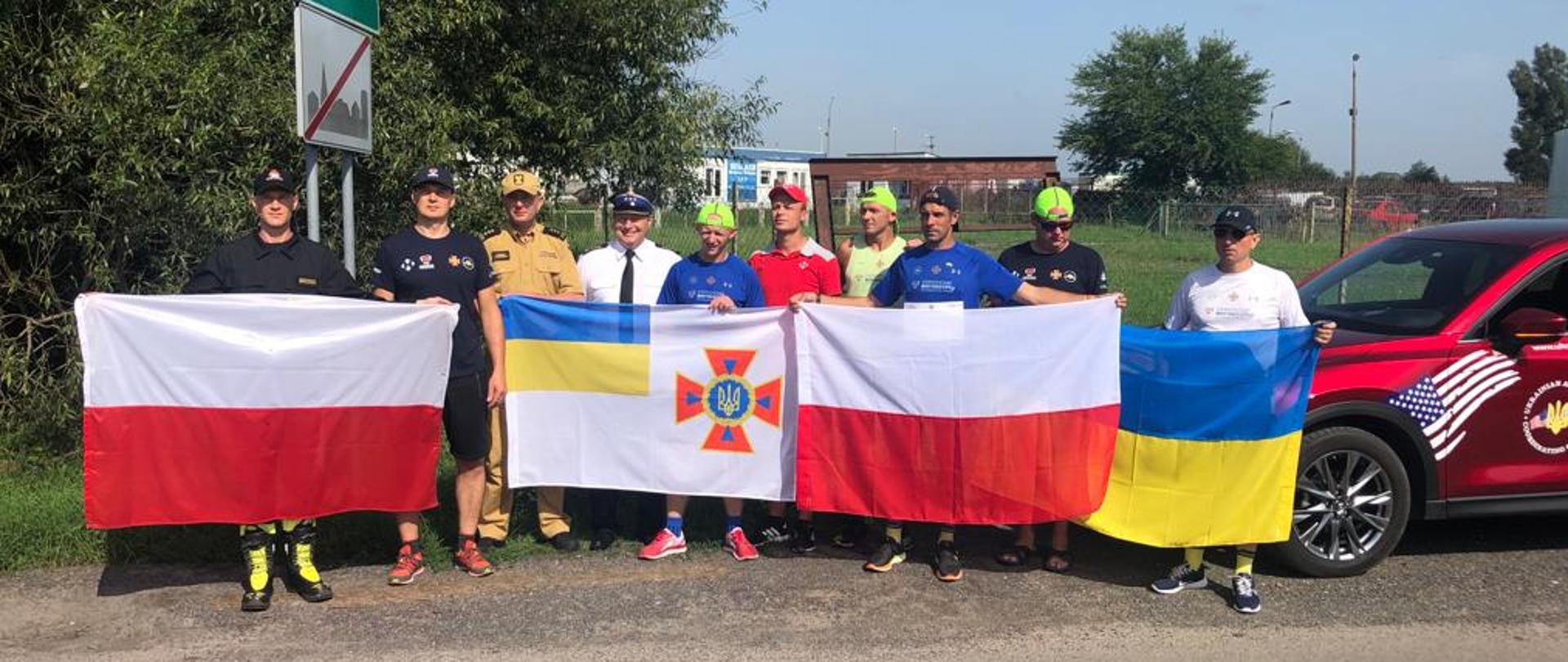 Trzech strażaków w mundurach oraz 7 w strojach sportowych pozują do zdjęcia na terenie otwartym trzymając flagi (od lewej) flafa Polski, Ukrainy z symbolem Państwowej Służby Ukrainy ds. Sytuacji Nadzwyczajnych, Polski i Ukrainy