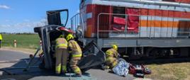 Na zdjęciu widać miejsce wypadku na przejeździe kolejowym: pociąg zatrzymał się przy uszkodzonym samochodzie, a wokół pracują strażacy i służby ratunkowe.