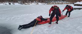 Zdjęcie przedstawia przemyskich strażaków podczas ćwiczenia z zakresu ratownictwa wodno-lodowego , trzech strażaków w skafandrach suchych i kaskach ochronnych w trakcie przemieszczania się po tafli lodu z użyciem sań lodowych