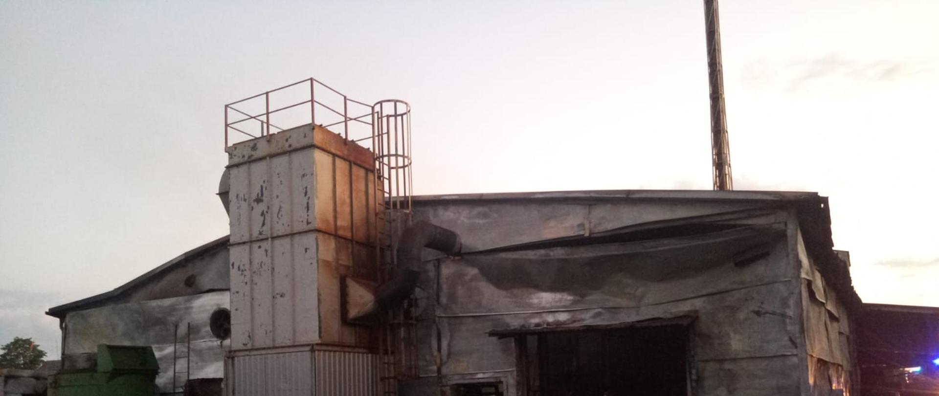 Widok tylnej ściany zniszczone, spalonej hali. W bramie do hali widoczni strażacy dogaszający elementy wyposażenia hali.