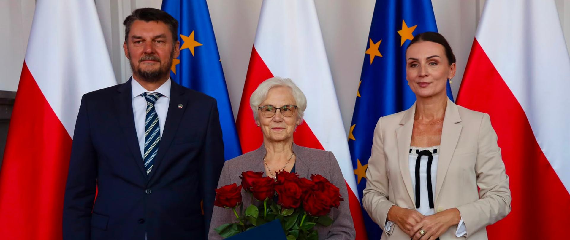 Na zdjęciu widoczna jest trójka osób pozujących w eleganckich strojach na tle naprzemiennie ustawionych flag Polski oraz Unii Europejskiej. Po lewej stronie stoi mężczyzna ubrany w granatowy garnitur, białą koszulę oraz krawat w niebiesko-białe paski. W centralnej części kadru znajduje się kobieta z jasnymi włosami w okularach, która trzyma w dłoniach bukiet czerwonych róż oraz niebieską teczkę ozdobioną godłem Polski. Po prawej stronie stoi kobieta ubrana w jasną marynarkę, ciemne spodnie oraz jasną bluzkę. Wszyscy uczestnicy wydarzenia patrzą w stronę aparatu.