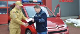 Komendant KP PSP w Brzezinach przekazuje dokumenty samochodu Citroen Berlingo prezesowi OSP Jordanów na tle samochodu oraz budynku komendy powiatowej PSP w Brzezinach