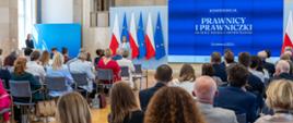 Wiceminister Paulina Piechna-Więckiewicz stoi przy mównicy i przemawia do ludzi siedzących na sali. W tle widać flagi Polski i Unii Europejskiej a na ekranie wyświetla się napis z tytułem konferencji.