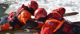 Na zdjęciu widoczni dwaj strażacy na skutym lodzie akwenie wodnym. Obaj w suchych skafandrach koloru pomarańczowego w czerwonych kaskach ochronnych. Jeden ratownik z użyciem drabin nasadkowych ewakuuje z wody drugiego strażaka.
