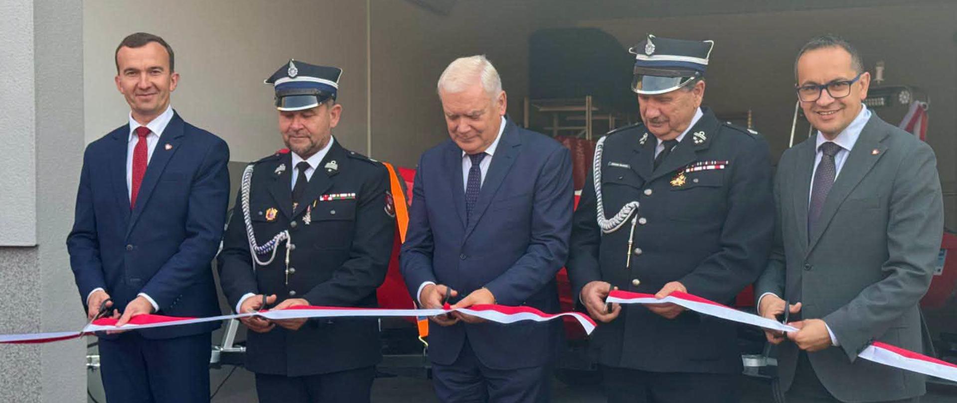 Grupa przedstawicieli władz, w tym wicewojewoda Mateusz Szauer, trzyma biało czerwoną wstęgę oraz nożyczki podczas uroczystego przecięcia wstęgi.
