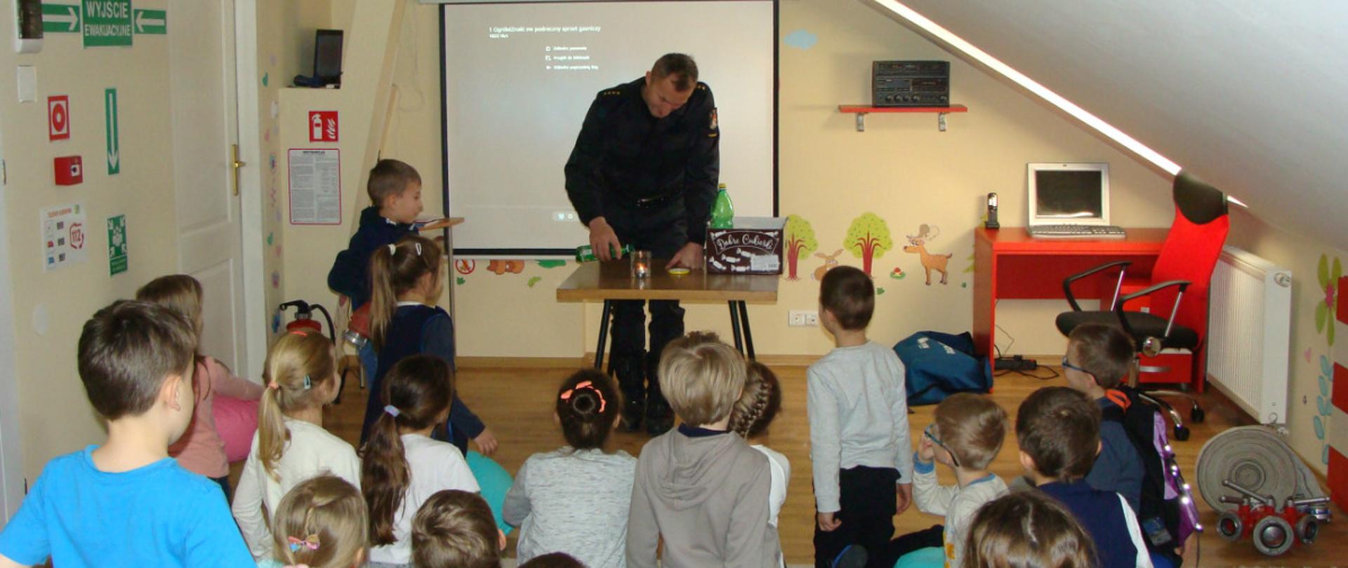 Dzieci w sali edukacyjnej "Ognik" z funkcjonariuszem prowadzącym zajęcia. 