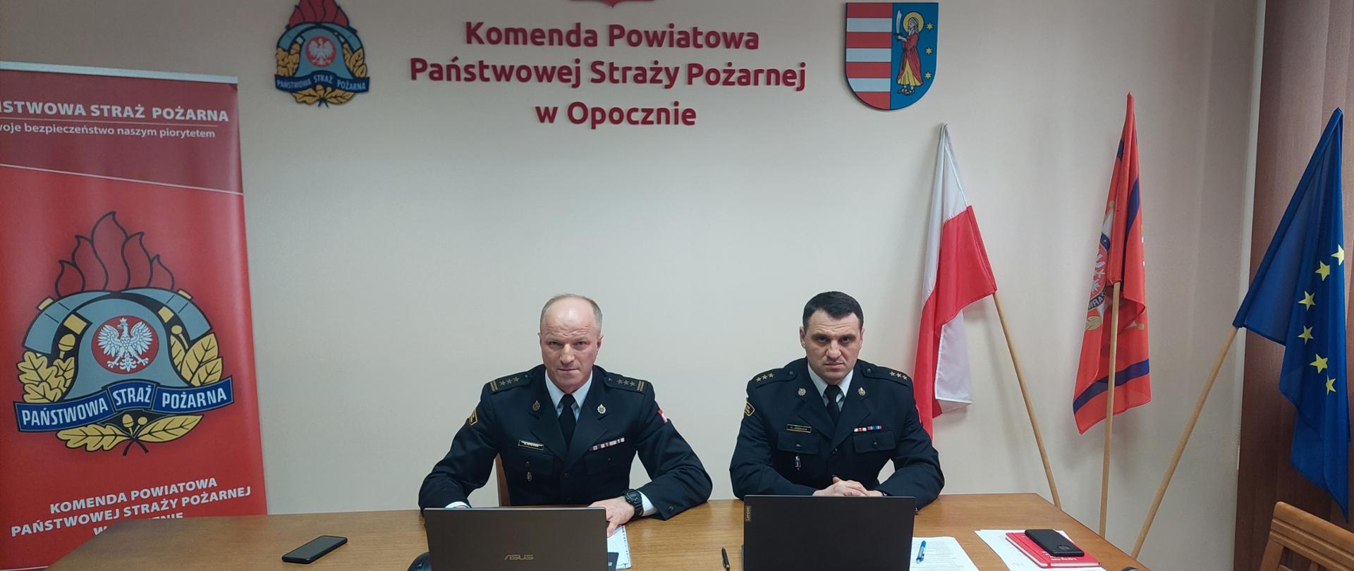Zdjęcie przedstawia Komendanta Powiatowego PSP w Opocznie i Jego zastępcę w mundurach wyjściowych koloru granatowego prowadzących wideokonferencję przy użyciu komputerów przenośnych. W tle znajdują się flagi koloru biało-czerwonego i niebieskiego oraz godło Polski, logo Państwowej Straży Pożarnej i logo Powiatu Opoczyńskiego.