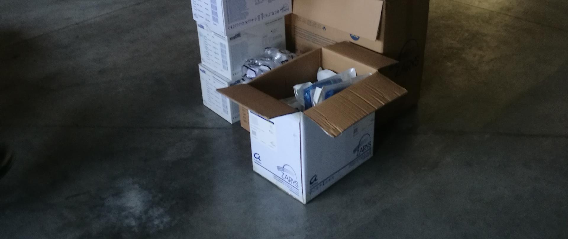 W garażu z betonową wylewką widać poukładane kartony, w których znajdują się środki ochrony indywidualnej przekazane dla PSP w Mławie. Na drugim planie pod ścianą na paletach poukładane jest więcej kartonów.