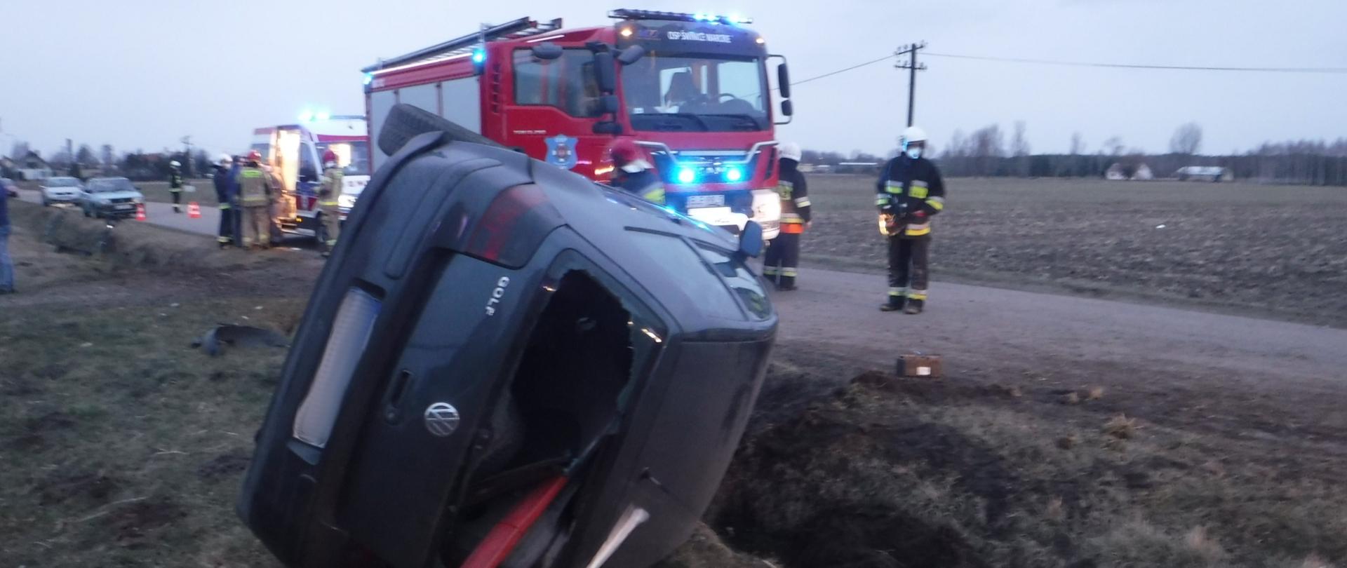 Na zdjęciu widać przewrócony na bok samochód osobowy, który znajduje się w rowie. W tle widać samochód strażacki, strażaków, karetkę pogotowia oraz stojące na poboczu samochody osobowe.