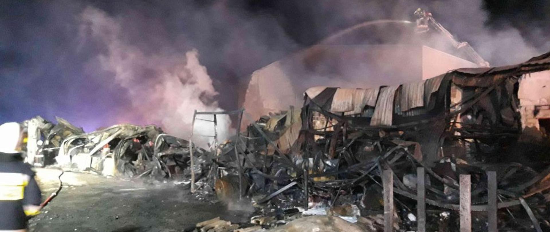 Na zdjęciu całkowicie spalona hala produkcyjna. Konstrukcyjne metalowe elementy całkowicie powyginane na skutek działania wysokiej temperatury. Z nad budynku unosi się dym. W tle drabina z samochodu strażackiego leje wodę na palącą się halę. Po lewej stronie strażak w trakcie działań. Zdjęcie wykonane w nocy.