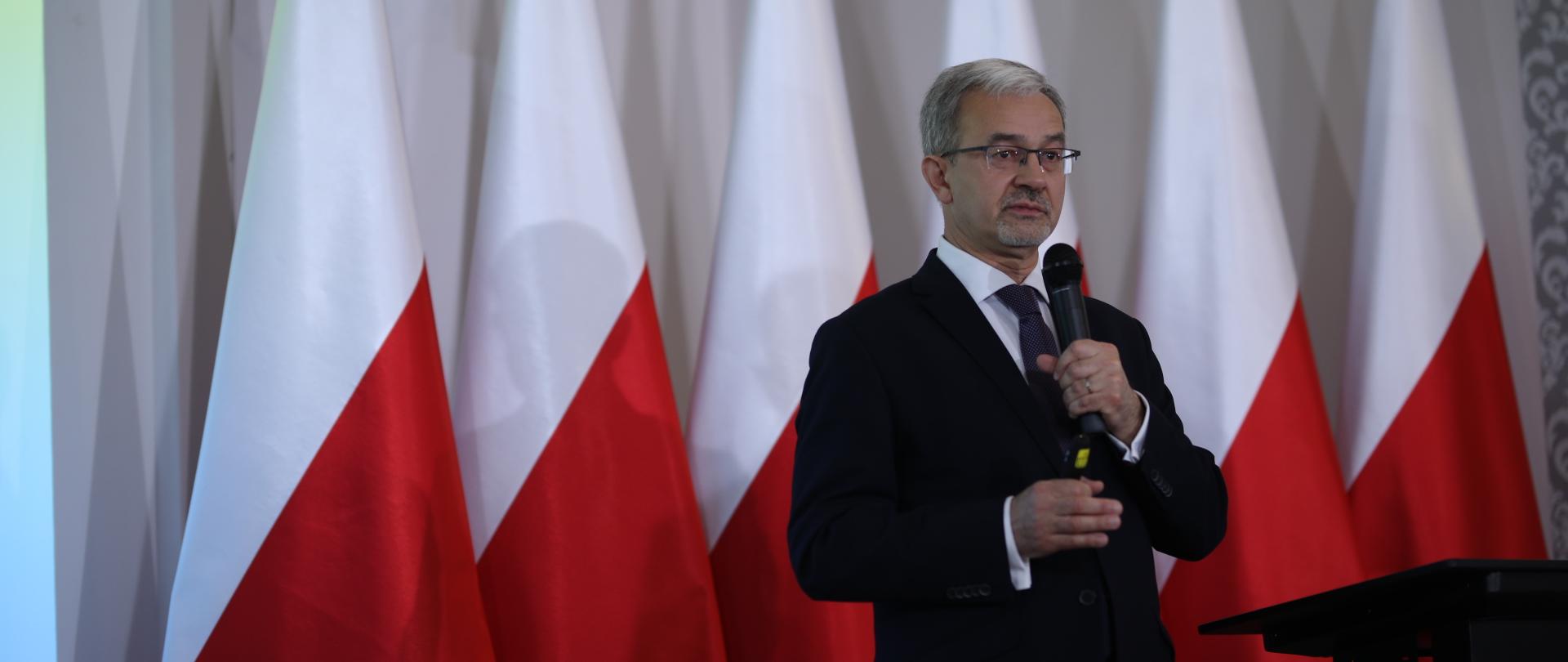 Na zdjęciu minister Jerzy Kwieciński z mikrofonem podczas przemówienia, w tle biało-czerwone flagi.