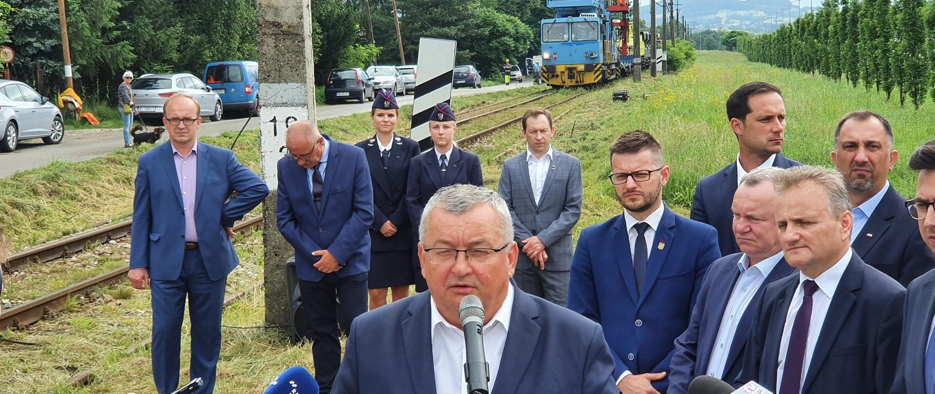 Na zdjęciu widać grupę ludzi, którzy stoją przy torach kolejowych. Na linii kolejowej widać prowadzone prace budowlane. Jeden z mężczyzn wypowiada się do mikrofonów.