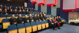 Na sali widowiskowej siedzą zaproszeni goście, strażacy ubrani są w mundury, w tle czerwone podświetlenia paneli