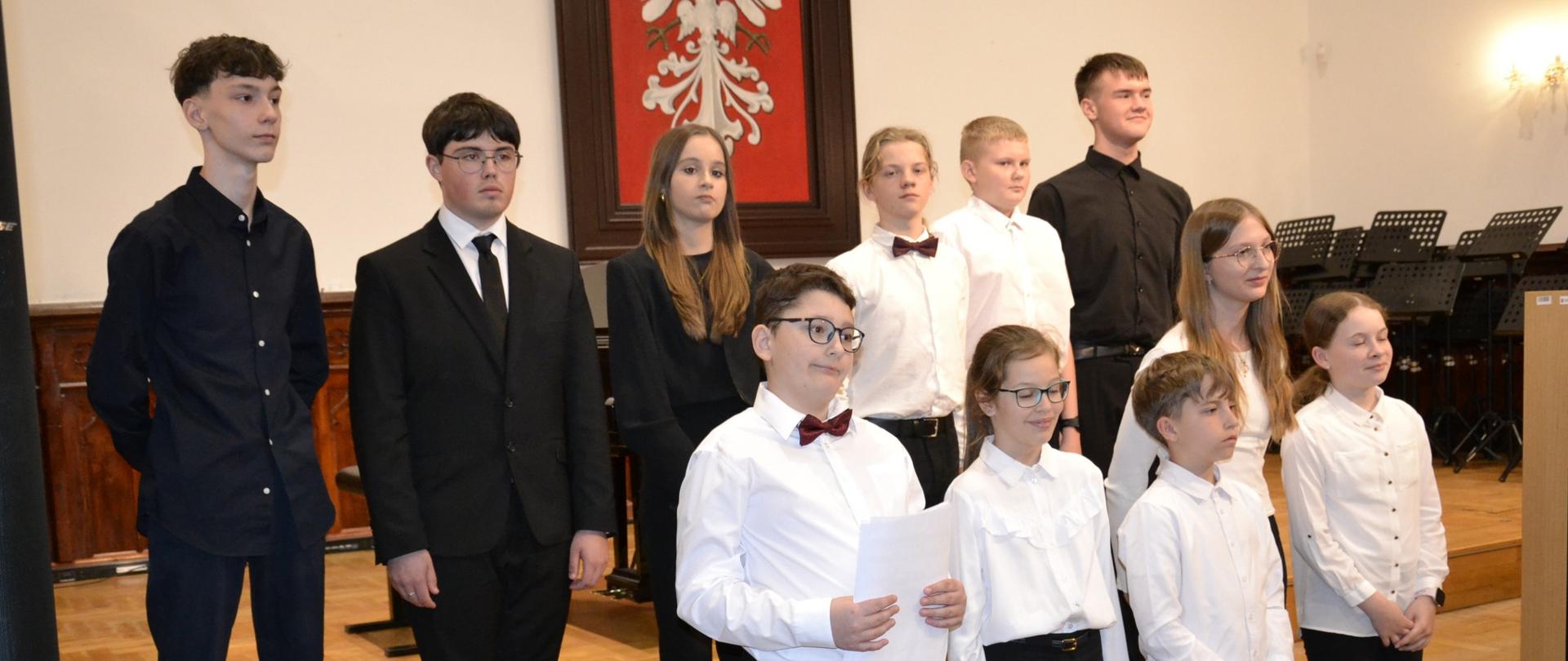Grupa uczniów w eleganckich strojach stoi na schodkach sceny Sali Królewskiej podczas koncertu klasy organów, w tle herb Polski.