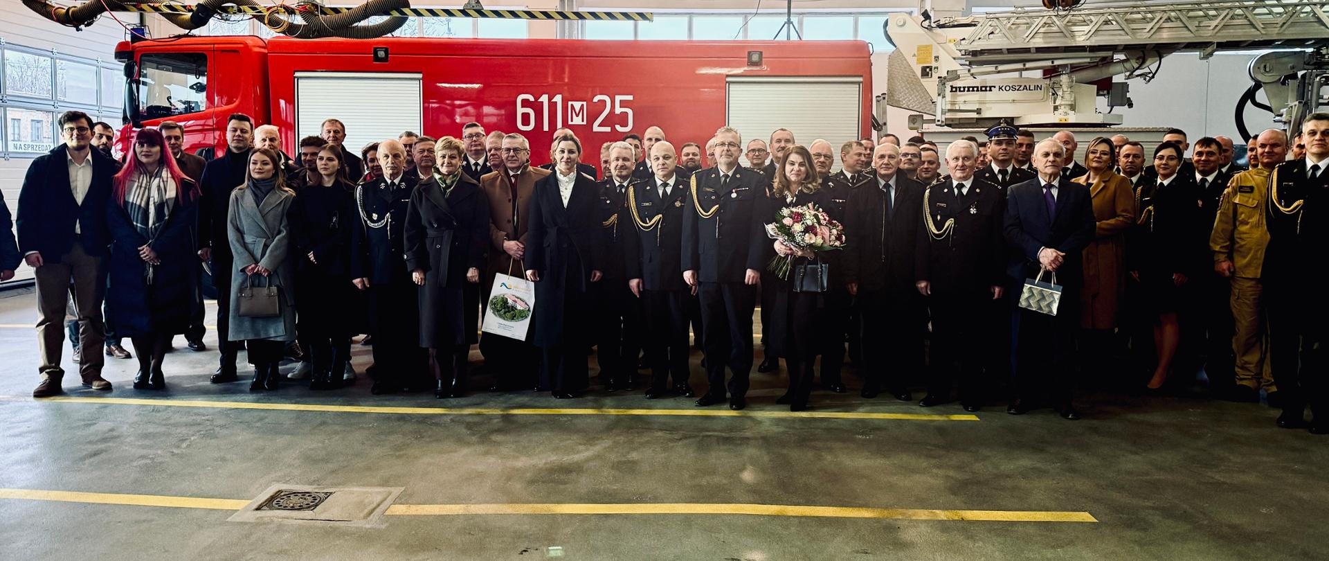 Na pierwszym planie stoi grupa ludzi obok siebie, kobieta trzyma kwiaty, a na drugim czerwone auto pożarnicze i z lewej strony widać bramę garażową.