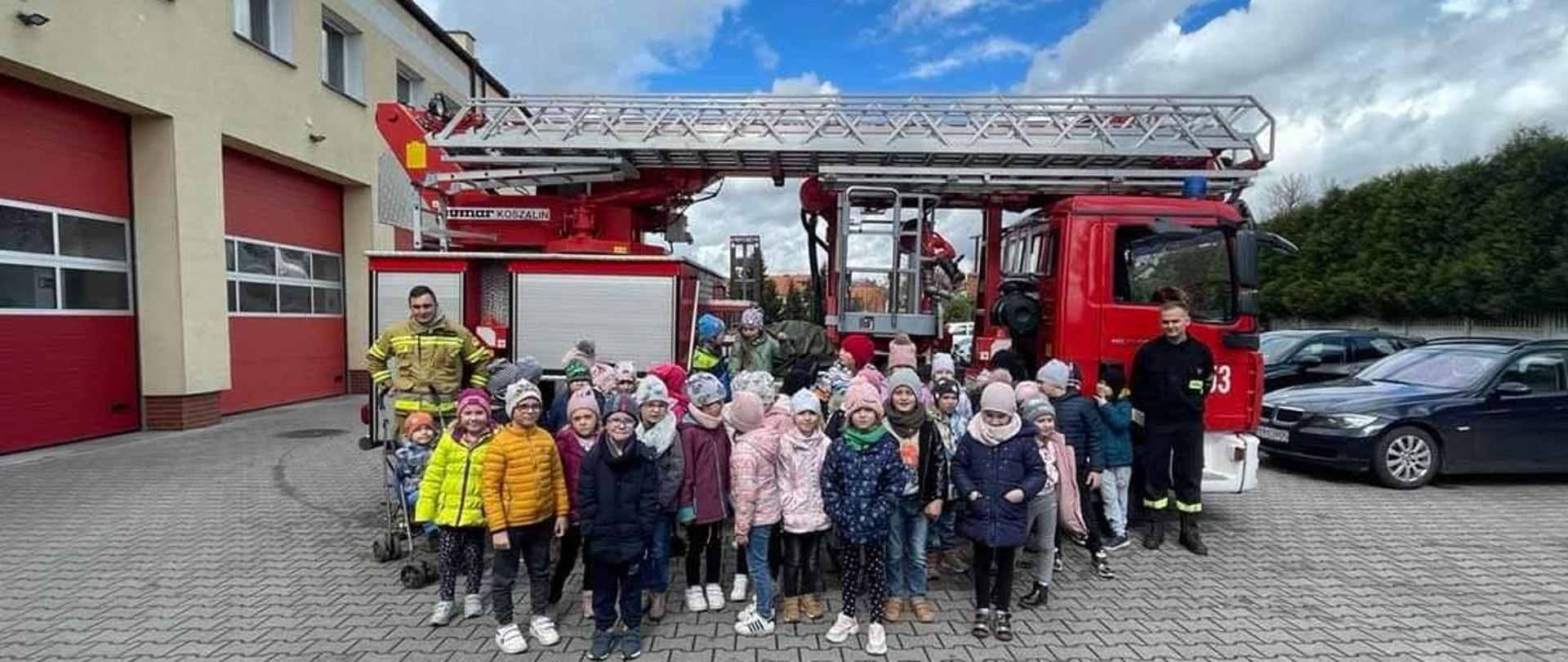 Na tle podnośnika hydraulicznego ustawionego na placu komendy dzieci wraz ze strażakami pozują do zdjęcia grupowego. 