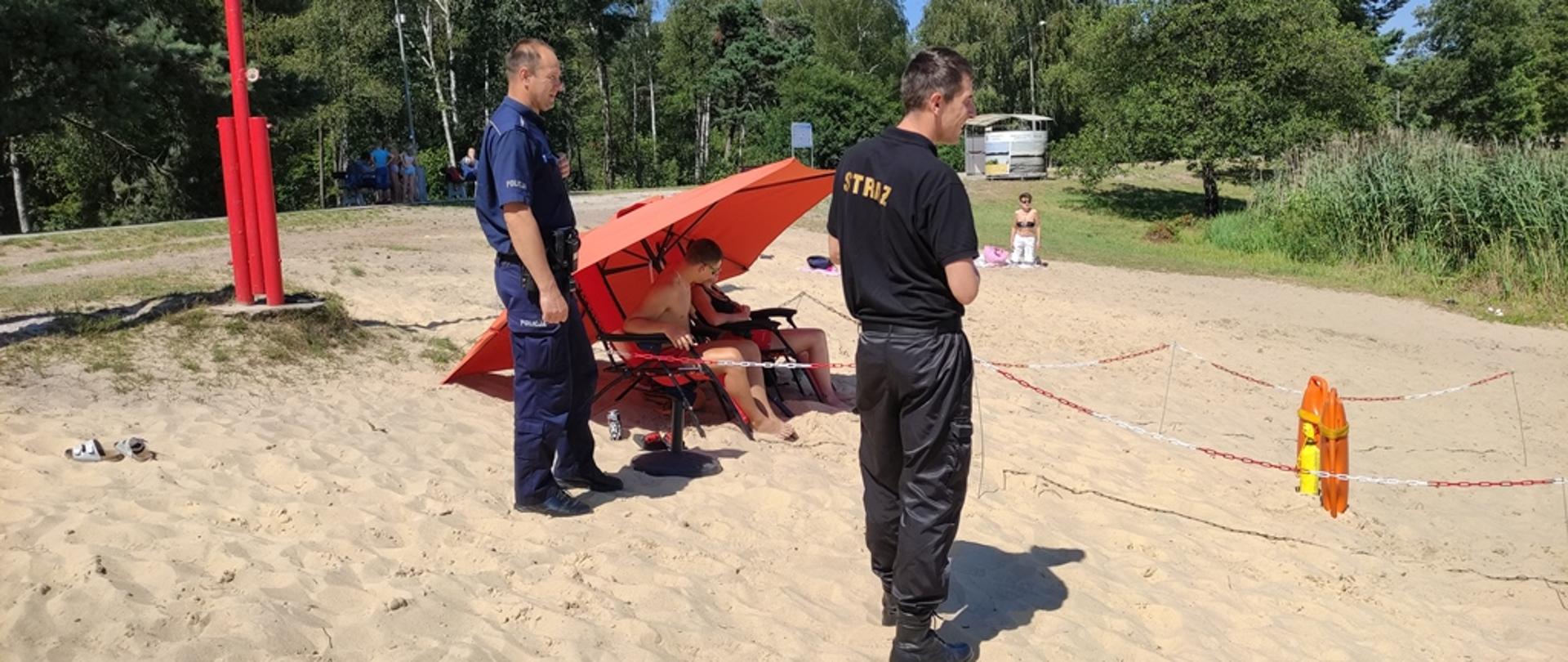 Teren zbiornika wodnego Klekot, plaża rejon kąpieliska, strażak wraz z policjantem stoją obok miejsca usytuowania ratowników wodnych czuwających nad bezpiecznym wypoczynkiem nad wodą, przed ratownikami w ziemię wbite boje oraz rzutka ratownicza 