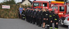 Strażacy OSP stoją w dwuszeregu, za nimi pojazdy pożarnicze, czekają na rozpoczęcie uroczystości.