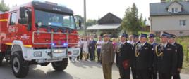 Na zdjęciu widzimy po lewej średni samochód ratowniczo-gaśniczy, a po prawej stojących w dwuszeregu funkcjonariuszy wojska, policji i straży pożarnej. W tle widoczni są zebrani goście oraz zabudowania.