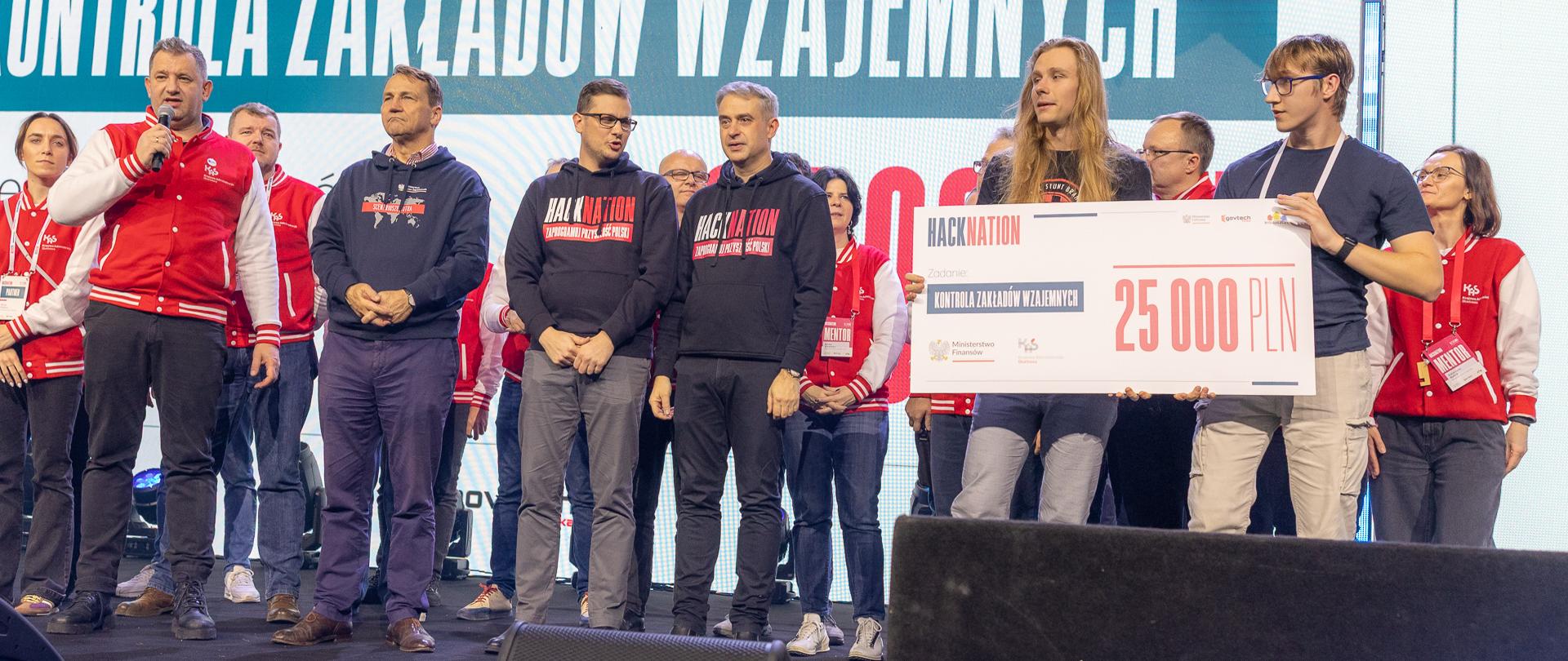Zespół EXTEMPORE odbiera nagrodę za wyzwanie „Kontrola zakładów wzajemnych” podczas HackNation 2025 w Bydgoszczy. Na scenie stoją przedstawiciele organizatorów i partnerów strategicznych wydarzenia, w tym Szef Krajowej Administracji Skarbowej oraz wicepremierzy Radosław Sikorski i Krzysztof Gawkowski. W tle widoczny duży ekran z nazwą wyzwania.