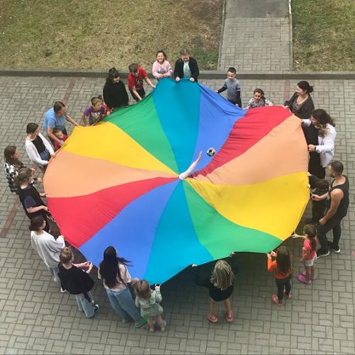 Widok z góry. Grupa dzieci stoi i trzyma wielką, kolorowa okrągłą chustę. Ze środka chusty wystaje ręka.