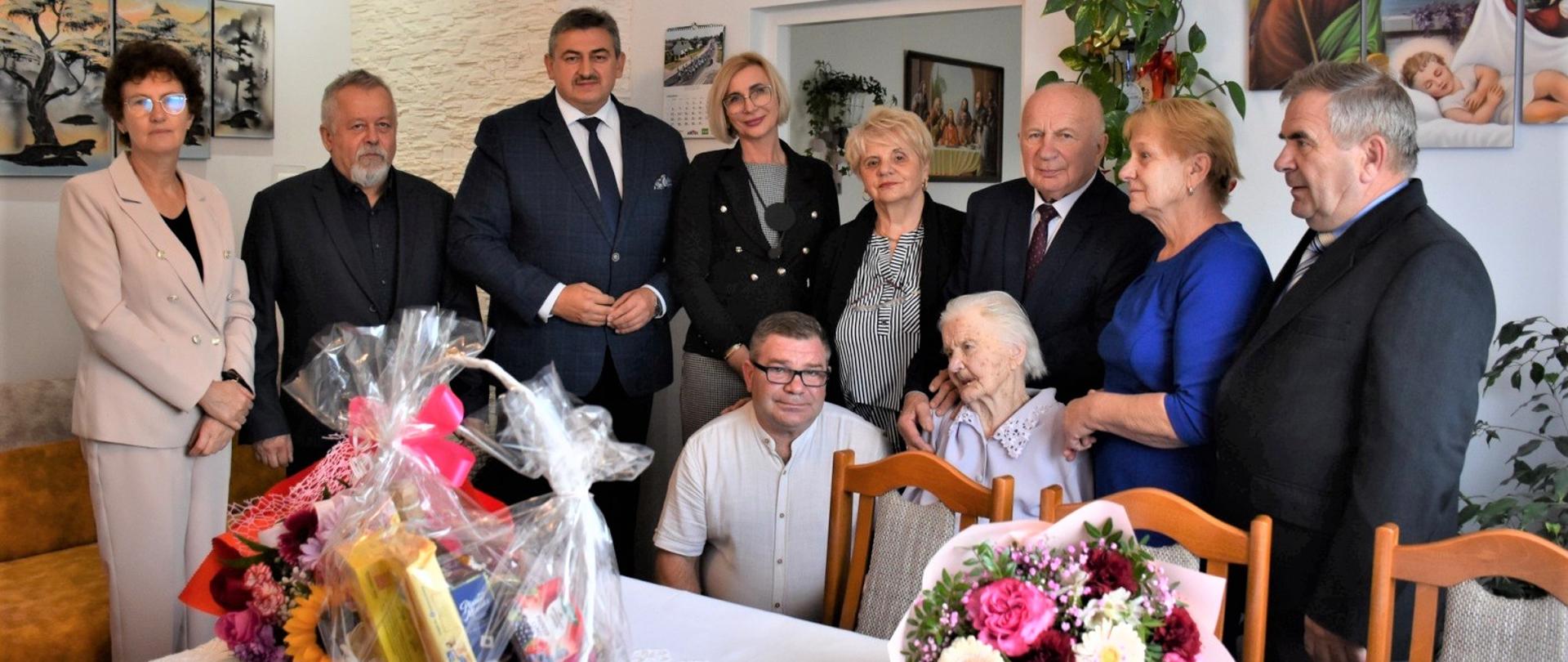 W pokoju pozuje do zdjęcia grupa osób – cztery kobiety i czterech mężczyzn stoją przy Jubilatce. Jubilatka siedzi na krześle. Po jej lewej stronie siedzi na krześle piąty mężczyzna. Przed nimi znajduje się stół, na którym znajdują się kwiaty oraz upominki.