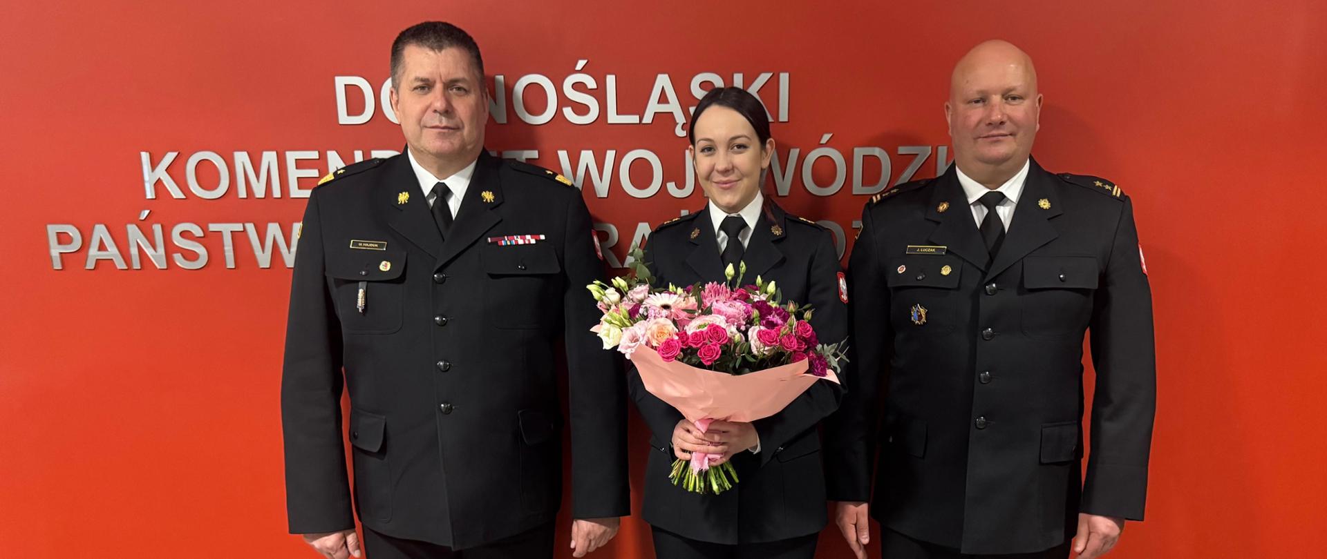 Zdjęcie przedstawia troje funkcjonariuszy Państwowej Straży Pożarnej w galowych mundurach, pozujących na tle czerwonej ściany z oficjalnym napisem. Centralna postać, kobieta, trzyma duży bukiet kwiatów.