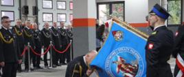 Komendant klęcząc na prawym kolanie całuje sztandar Komendy. Sztandarowi salutują. W tle inni strażacy w mundurach.