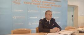 p.o. Komendant Szkoły Aspirantów PSP w Krakowie mł.bryg. Dariusz Ruchała - strażak w umundurowaniu wyjściowym siedzący na tle niebieskiej ścianki z logotypami Szkoły 