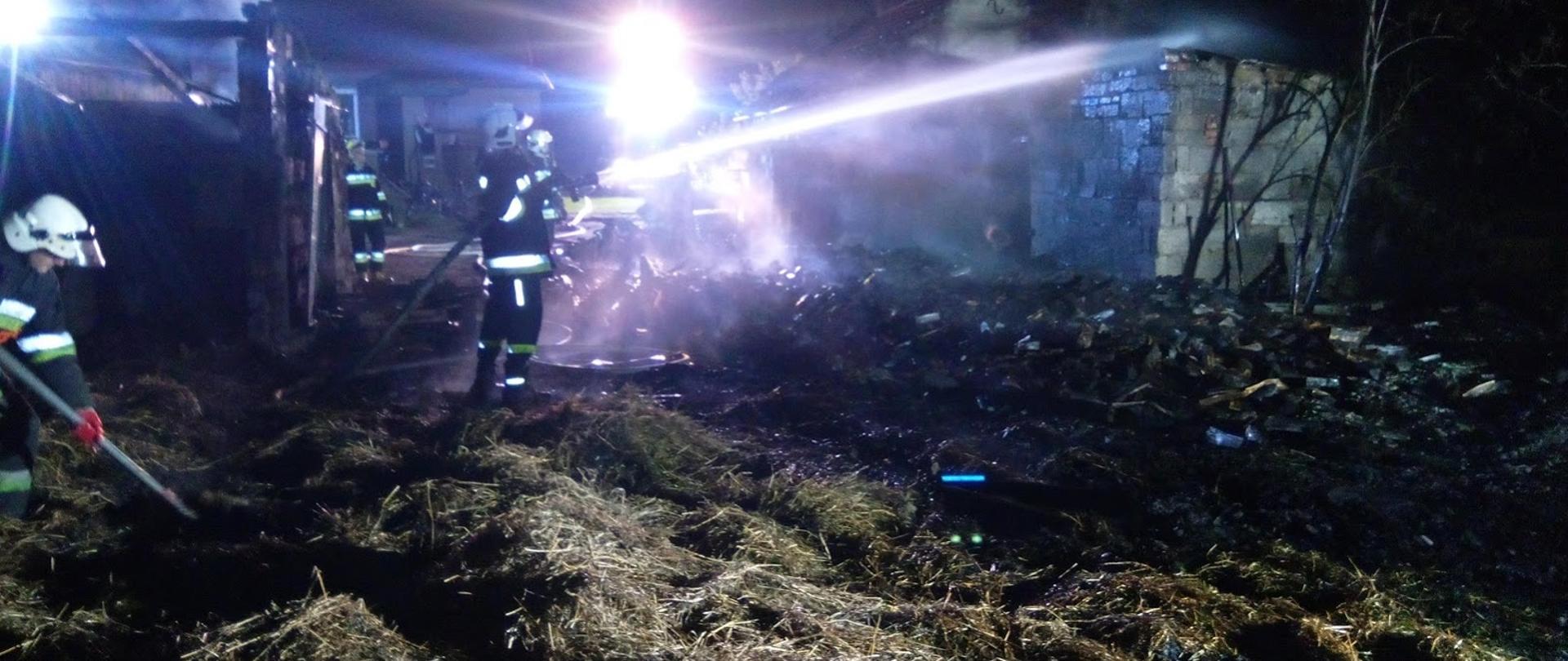 . Zdjęcie przedstawia pogorzelisko po pożarze budynku gospodarczego ze spalonymi elementami konstrukcji. Widoczny unoszący się dym, w tle strażak OSP dogaszający pogorzelisko prądem wody.