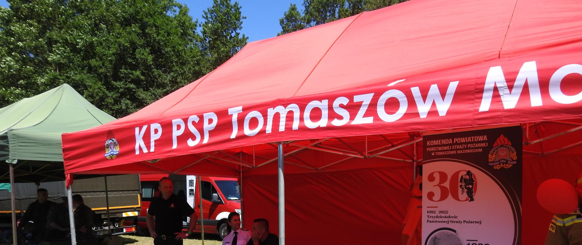Duży czerwony namiot z białym napisem KP PSP Tomaszów Maz. Z lewej strony pod namiotem, przy stoliku na którym rozłożone są ulotki, słodycze i inne materiały siedzą strażacy. Z prawej strony stoi baner reklamowy otoczony na dole czerwonymi balonami. Obok niego manekin ubrany w ubranie specjalne i hełm.