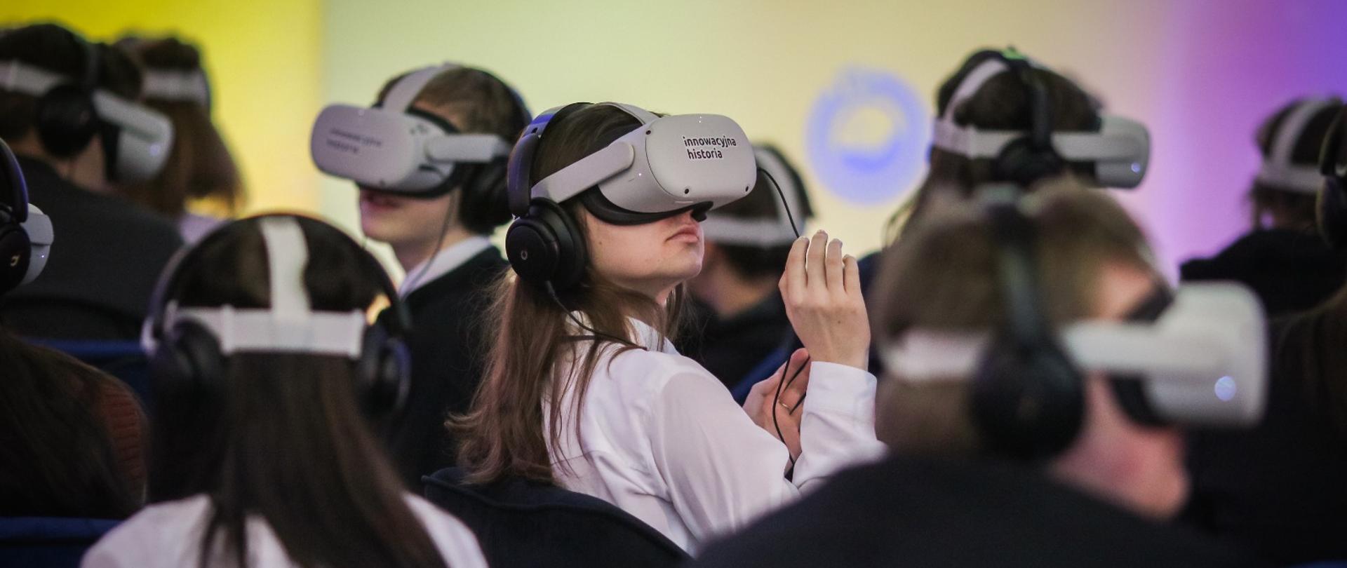W sali z kolorowymi ścianami siedzi wielu młodych ludzi, na głowach mają gogle do wirtualnej rzeczywistości.