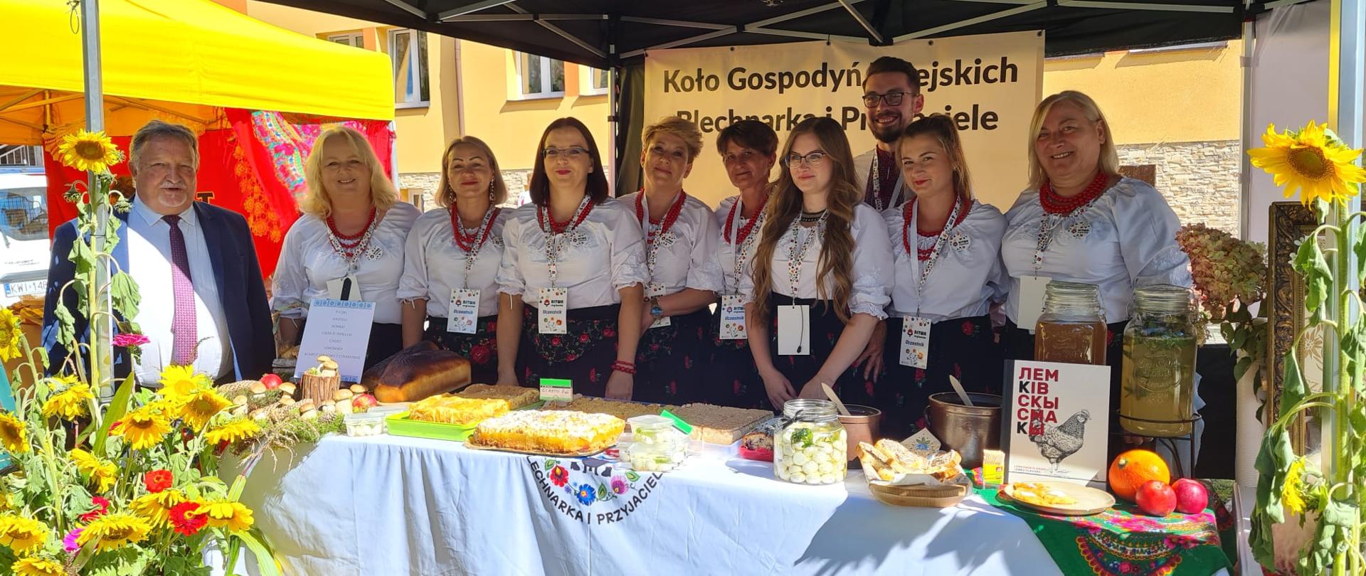 Grupa osób (kobiety i mężczyzna) za stoiskiem Koła Gospodyń Wiejskich „Blechnarka i Przyjaciele”. Na białym obrusie leżą chleby, ciasta i regionalne produkty. Obok stoi starszy mężczyzna w garniturze.