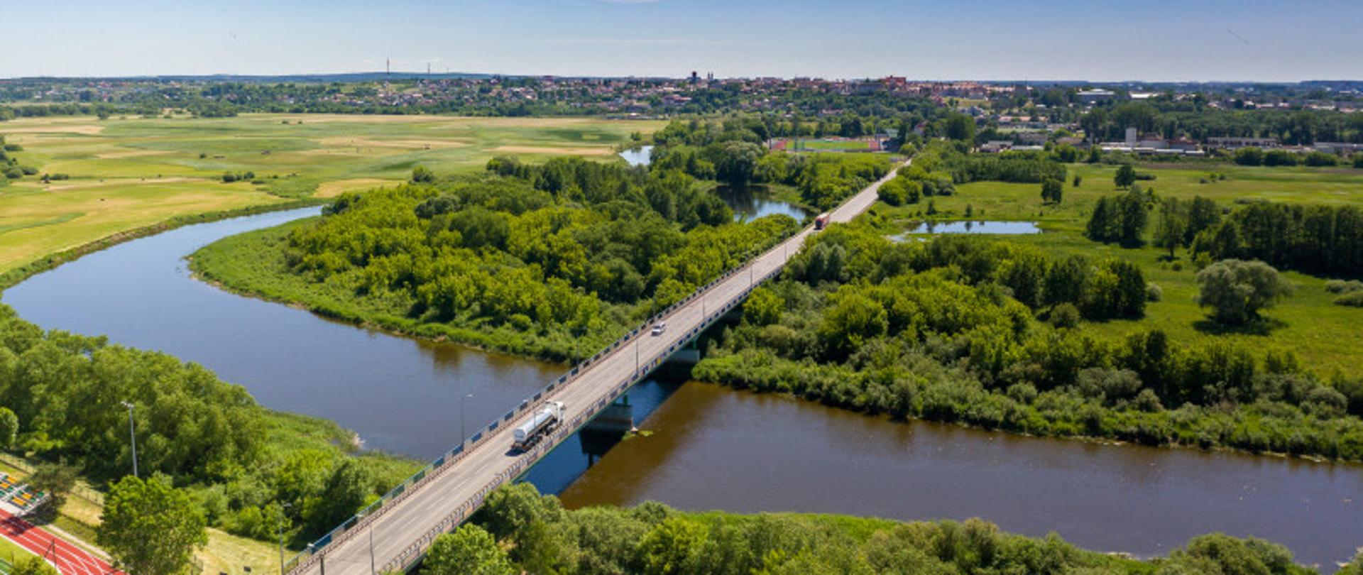 Zdjęcie przedstawia most nad rzeką, po którym jadą pojazdy. W tle drzewa, pola, a na dalszym planie widoczne zabudowania mieszkalne.
